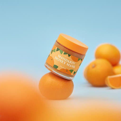 Jar of hydrating mandarin honey mask balanced on fresh mandarin with whole and sliced mandarins on soft blue background