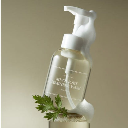 Clear pump bottle of soothing feminine wash with foamy lather and fresh green mugwort leaf on beige background
