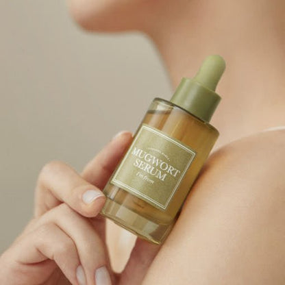 Hand holding a green dropper bottle of soothing mugwort serum against bare shoulder and neutral background