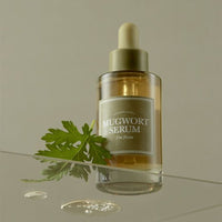 Clear glass dropper bottle of soothing mugwort serum with fresh green leaf and subtle liquid drops nearby