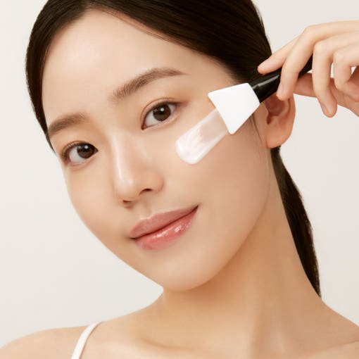 Young woman applying white peel off mask with silicone brush, demonstrating elasticity-boosting skincare treatment.