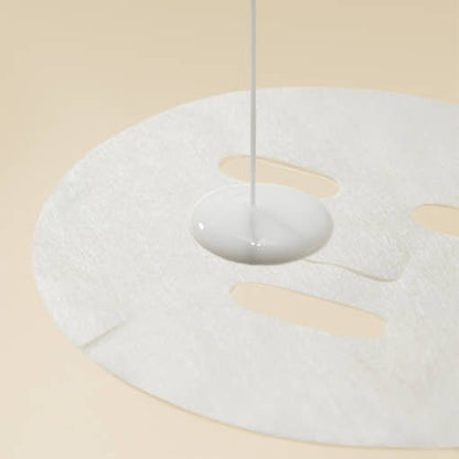 White sheet mask with hydrating liquid being poured onto smooth beige background surface
