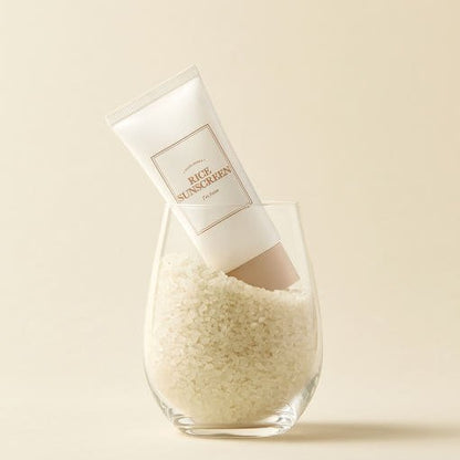 White sunscreen tube labeled rice sunscreen standing in a clear glass filled with white rice grains on beige background