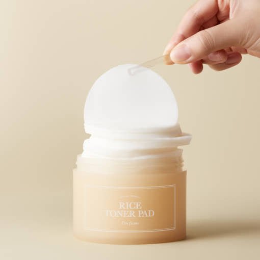 Hand removing a round toner pad from a beige container with a clear spatula on neutral background