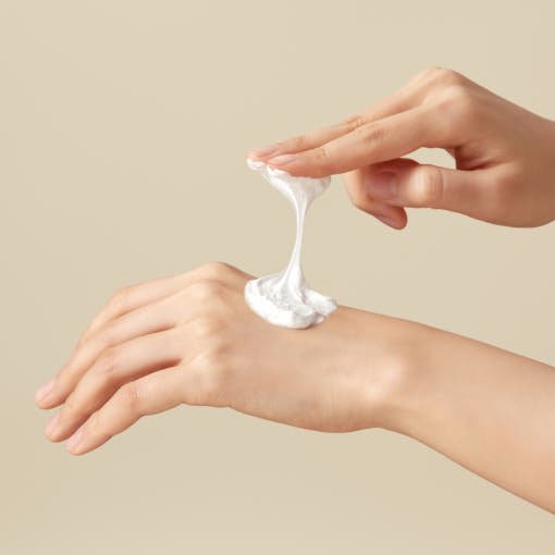 Creamy white foam being gently stretched between fingers above the back of a hand against beige background