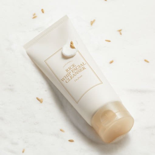 Creamy white cleansing foam on a white tube with gold cap and scattered rice grains on light background