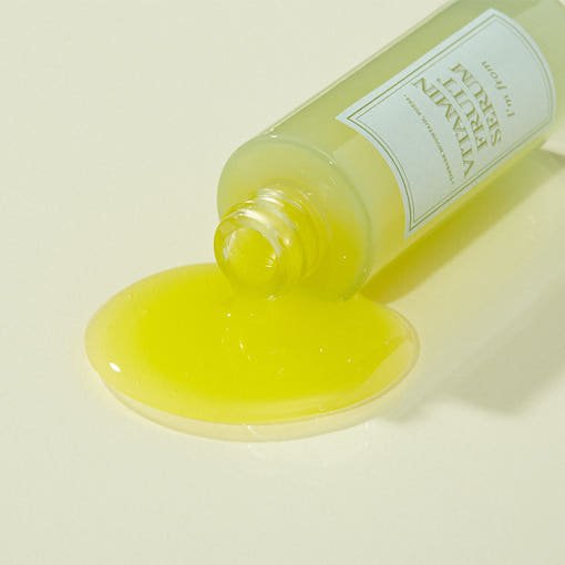 Yellow vitamin serum spilling from transparent dropper bottle on pale yellow surface with soft shadows