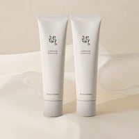 Two minimalist white tubes of hydrating cream with subtle black and red text on soft beige fabric background
