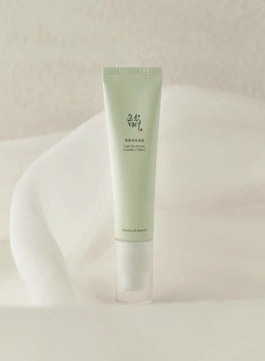Pale green tube of brightening serum with simple black Korean text standing on soft white fabric background