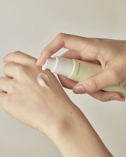 Creamy serum being dispensed from light green pump bottle onto back of hand, showing smooth texture and brightening benefit