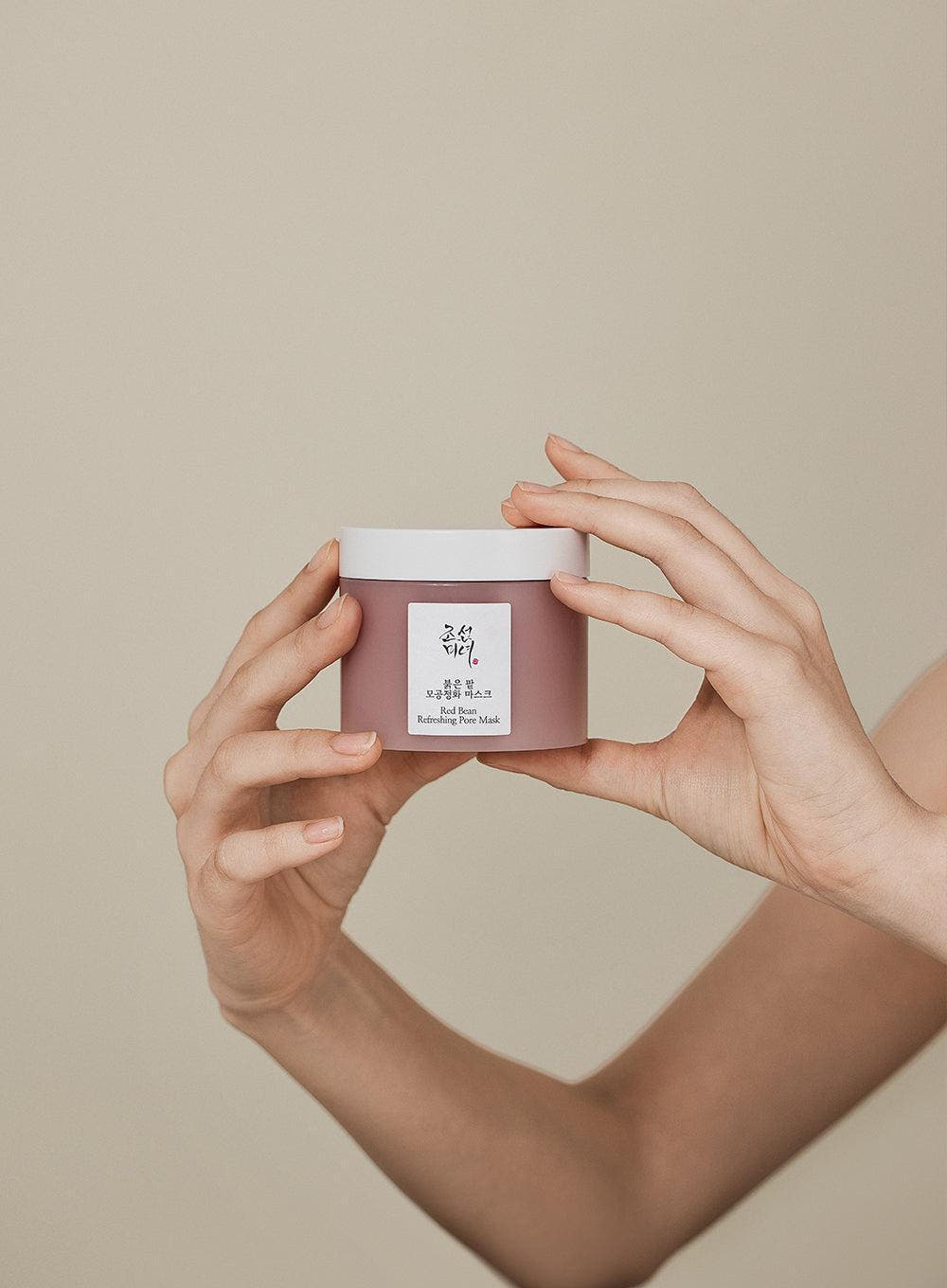 Hands holding a muted red jar of pore-cleansing mask with a clean white lid against a plain background