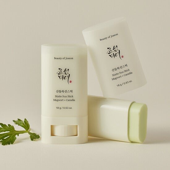 Two matte sun sticks with pale green balm and soft white packaging alongside green leaves on beige background