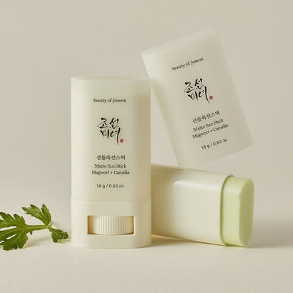 Two matte sun sticks with pale green balm and soft white packaging alongside green leaves on beige background