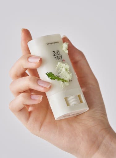 Hand holding a white sun stick with green herbal cream and a small leaf accent on a plain background
