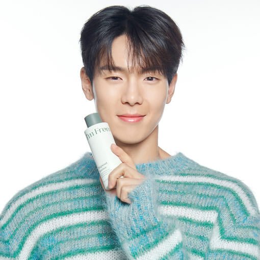 Young man in striped blue-green sweater holding a white bottle of hydrating mugwort essence against white background