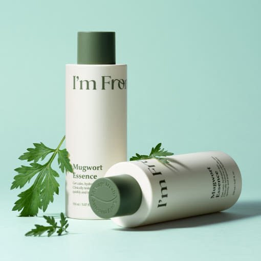 Two white cylindrical bottles with green caps labeled Mugwort Essence with fresh green leaves on mint background