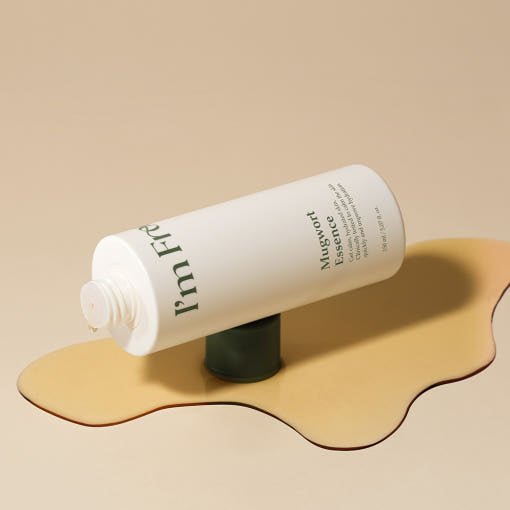White bottle of hydrating mugwort essence resting on its cap over a beige liquid spill on neutral background