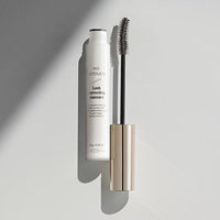 Open clear mascara tube with silver cap and curved brush on soft gray surface with shadow contrast