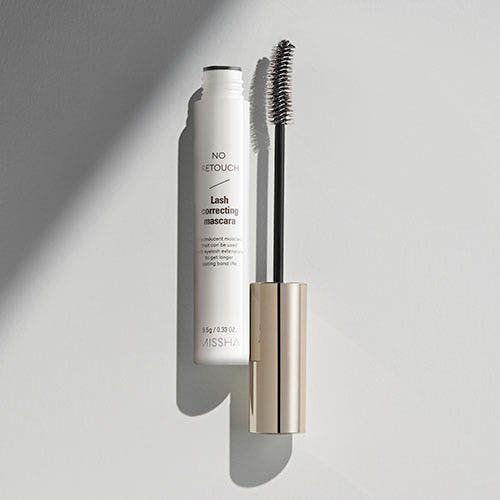 Open clear mascara tube with silver cap and curved brush on soft gray surface with shadow contrast