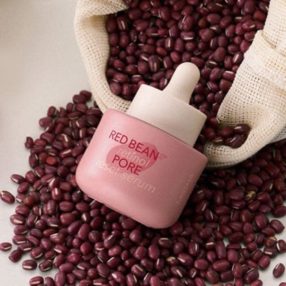Pink dropper bottle of pore minimizing serum surrounded by red beans spilling from a woven sack