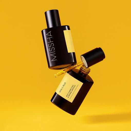 Two sleek black ampoule bottles with yellow labels, one open with a dropper releasing brightening serum against yellow background
