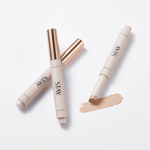 Beige concealer sticks with metallic rose gold caps and creamy high coverage formula swatch on white background