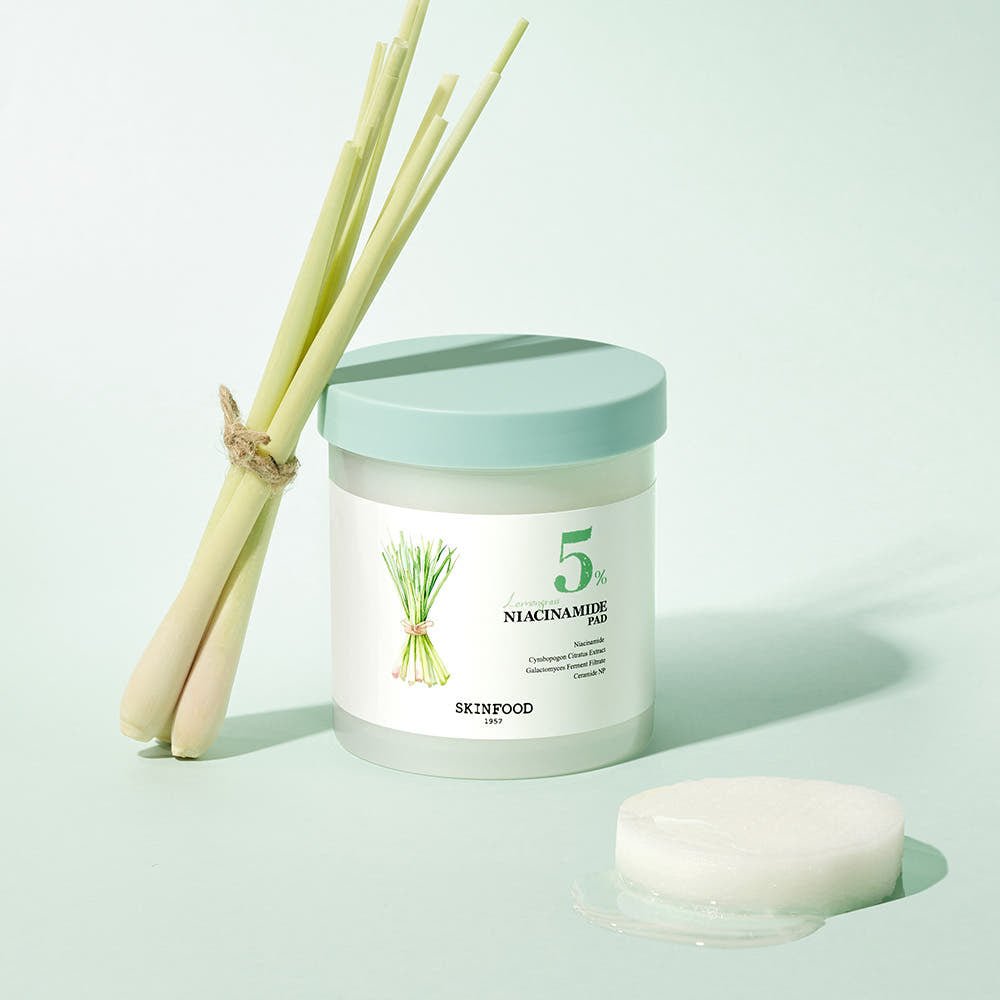 Jar of lemongrass-infused niacinamide pads with light green lid and lemongrass stalks on pale background