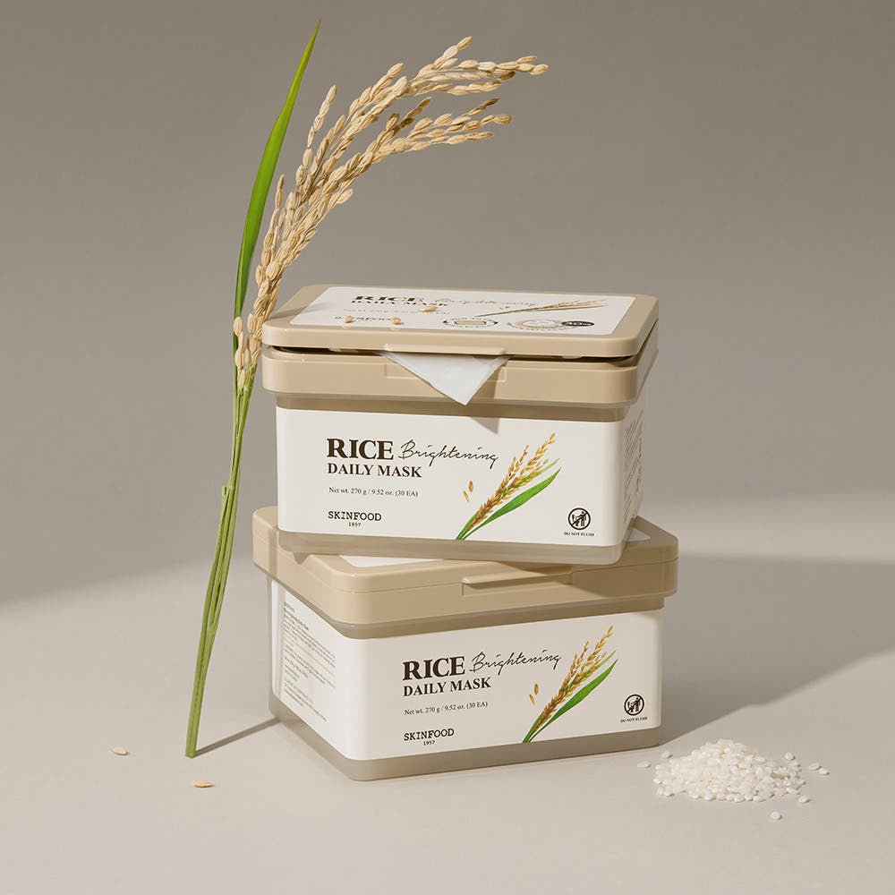 Two beige storage containers of rice brightening daily mask stacked beside rice grains and rice stalk on neutral background