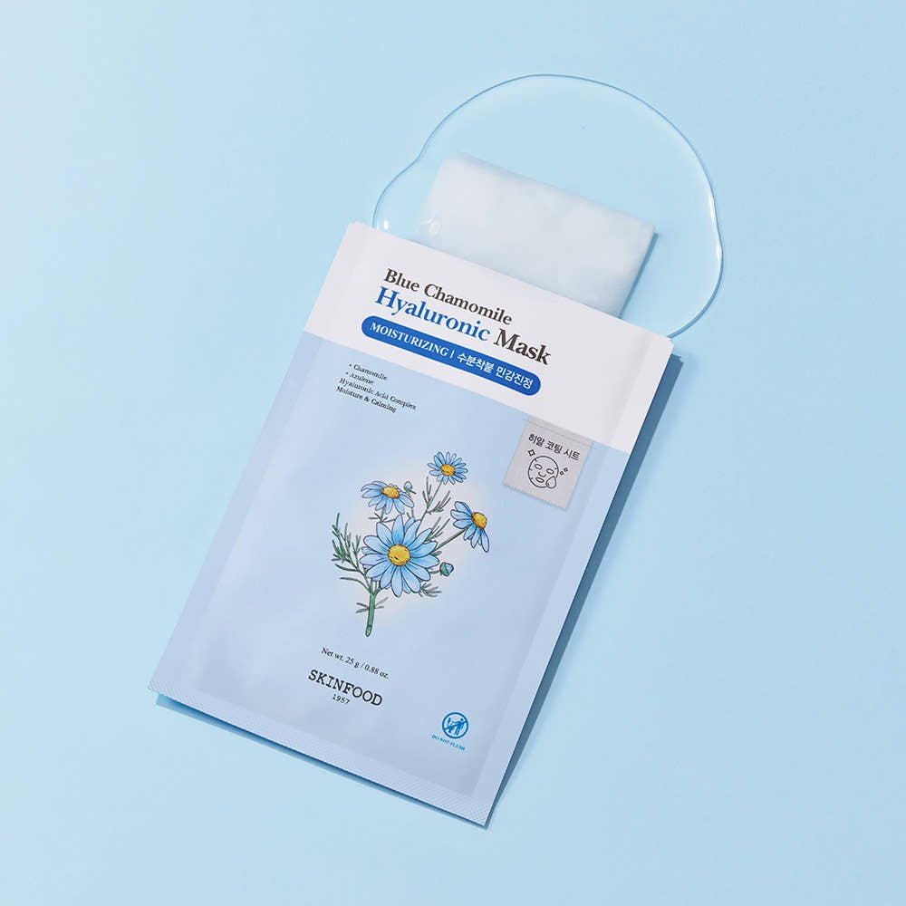 Light blue packet of hydrating facial sheet mask with chamomile flower illustration on blue surface with serum spilled