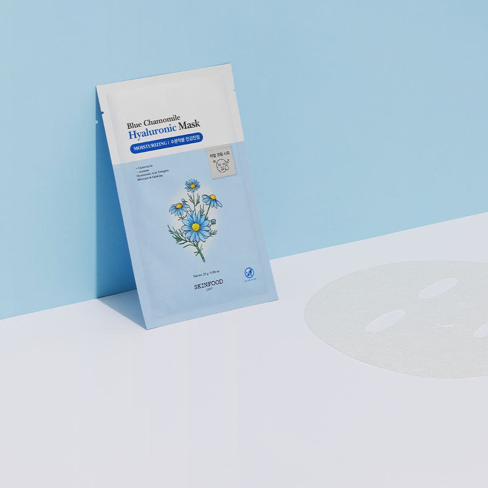 Blue chamomile hyaluronic mask sheet packet standing on pale blue and white surface next to unfolded translucent face mask sheet