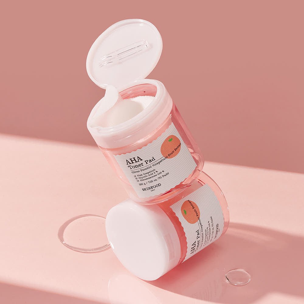 Two stacked translucent pink jars of gentle exfoliating AHA toner pads with peach fruit label on soft pink surface