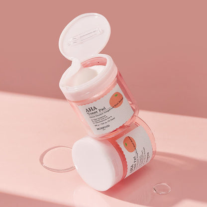 Two stacked translucent pink jars of gentle exfoliating AHA toner pads with peach fruit label on soft pink surface