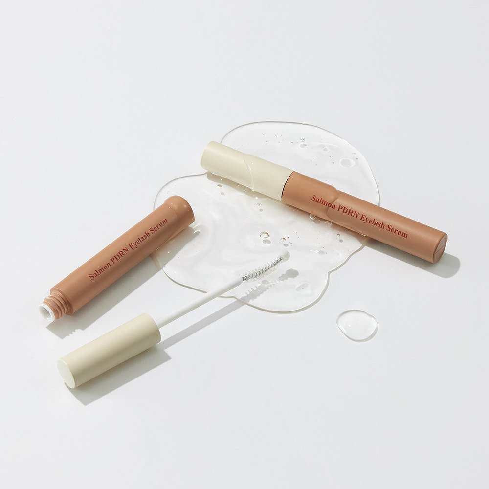 Two beige and cream eyelash serum tubes with wand applicator and clear serum spilled on white surface