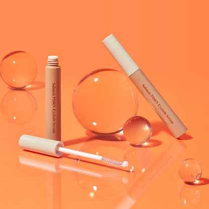 Two beige eyelash serum tubes with applicator wands and translucent spheres on glossy orange surface