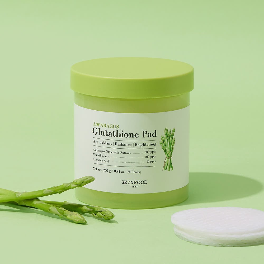 Green container of brightening glutathione pads with asparagus illustration next to fresh asparagus and cotton pads on green background