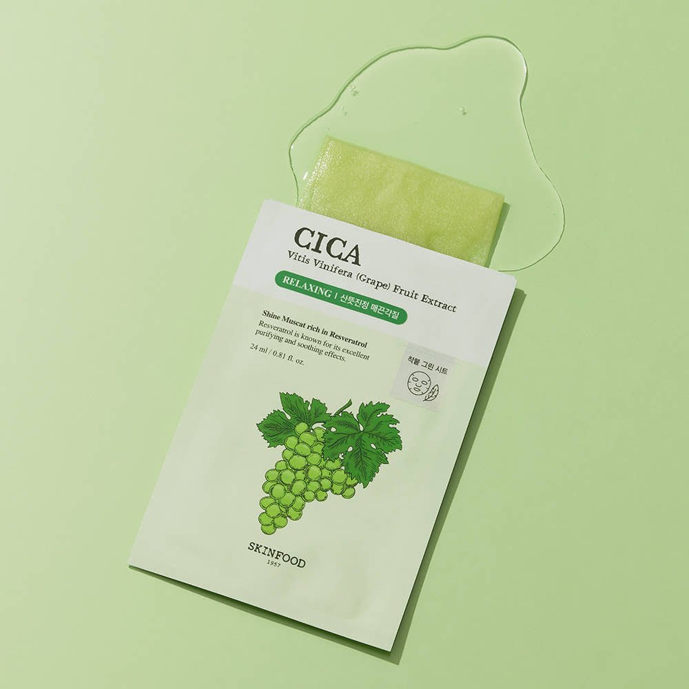 Light green sheet mask partially pulled out of white sachet featuring grape illustration and clear serum spill on pale green surface