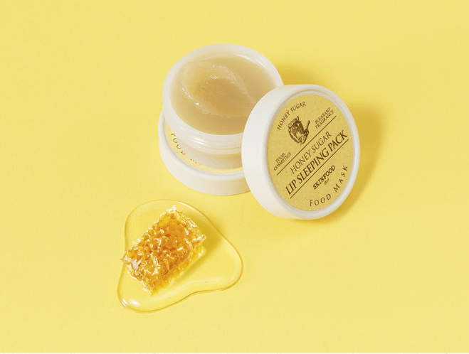 Open jar of honey sugar lip mask with smooth texture beside honeycomb piece and golden honey on yellow background