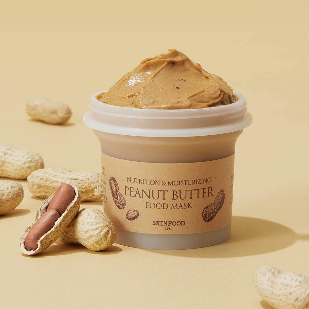 Open jar of peanut butter food mask with creamy texture surrounded by whole and shelled peanuts on beige surface
