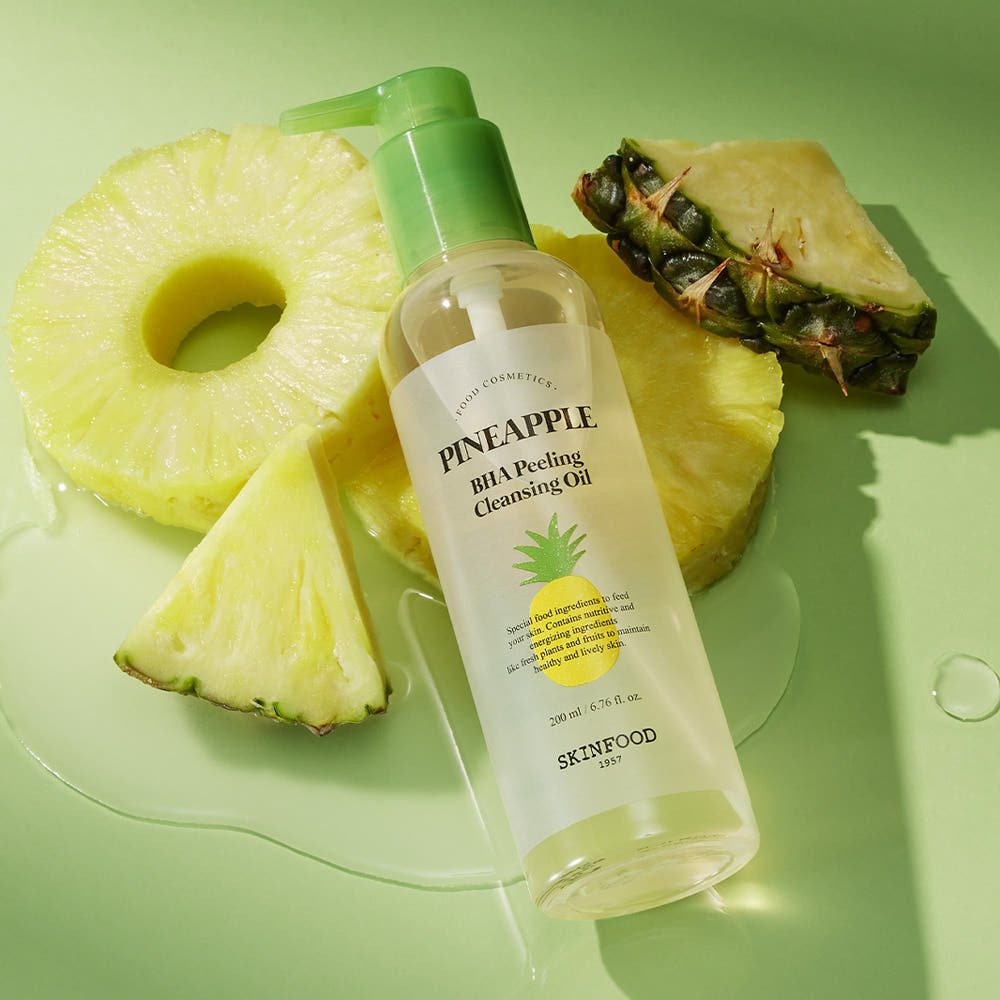 Clear bottle of pineapple-infused peeling cleansing oil with green pump surrounded by fresh pineapple slices on green surface