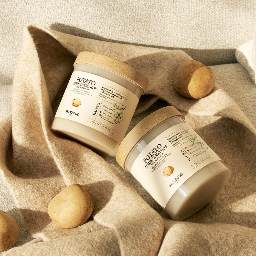 Two skincare containers with natural wood lids surrounded by small potatoes on soft beige fabric background