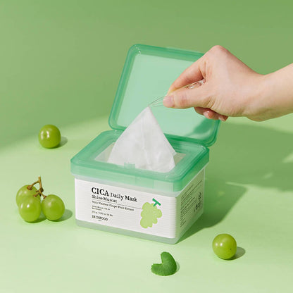 Hand lifting a sheet mask with tweezers from a green-topped container surrounded by Shine Muscat grapes on green background