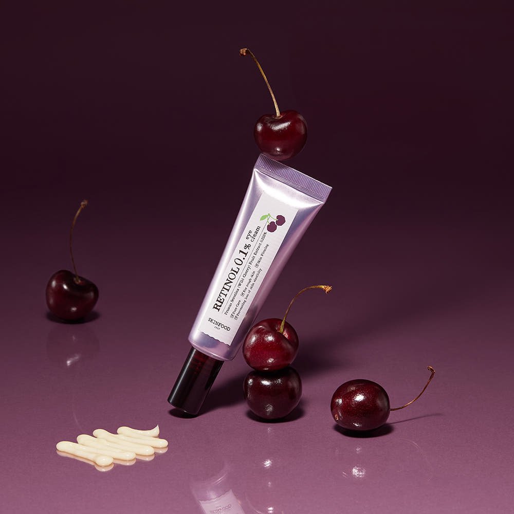 Purple tube of retinol eye cream standing among fresh cherries with cream swatch on glossy surface