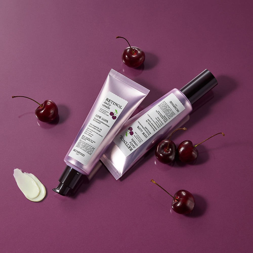 Two pink tubes of retinol cream with black caps beside fresh cherries and cream swatch on purple surface