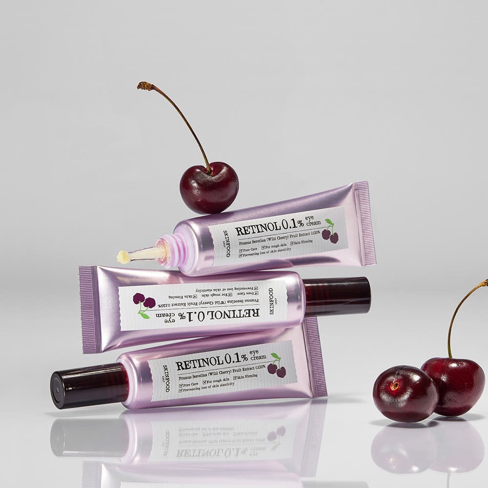 Three lavender-toned tubes of retinol eye cream surrounded by fresh dark cherries on a reflective surface
