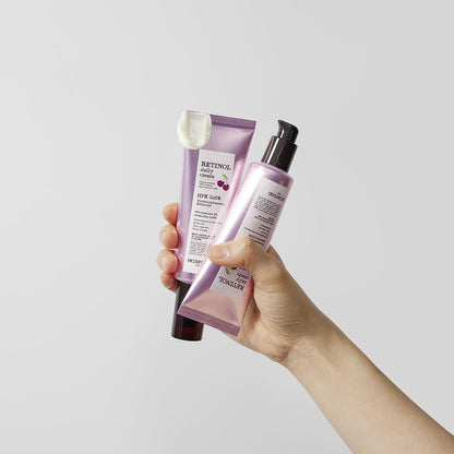 Hand holding purple tubes of retinol daily cream with smooth white cream texture on a pale background
