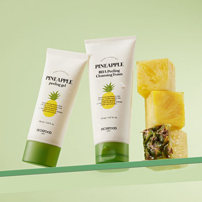 Two white tubes of pineapple exfoliating skincare products with green caps beside stacked fresh pineapple chunks on pale green background