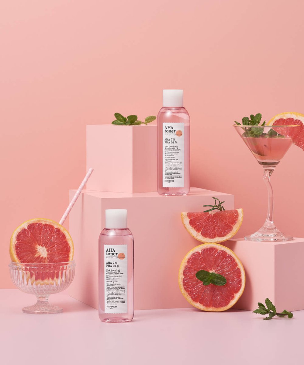 Two pink bottles of exfoliating AHA toner displayed with fresh pink grapefruit slices and mint leaves on pink blocks