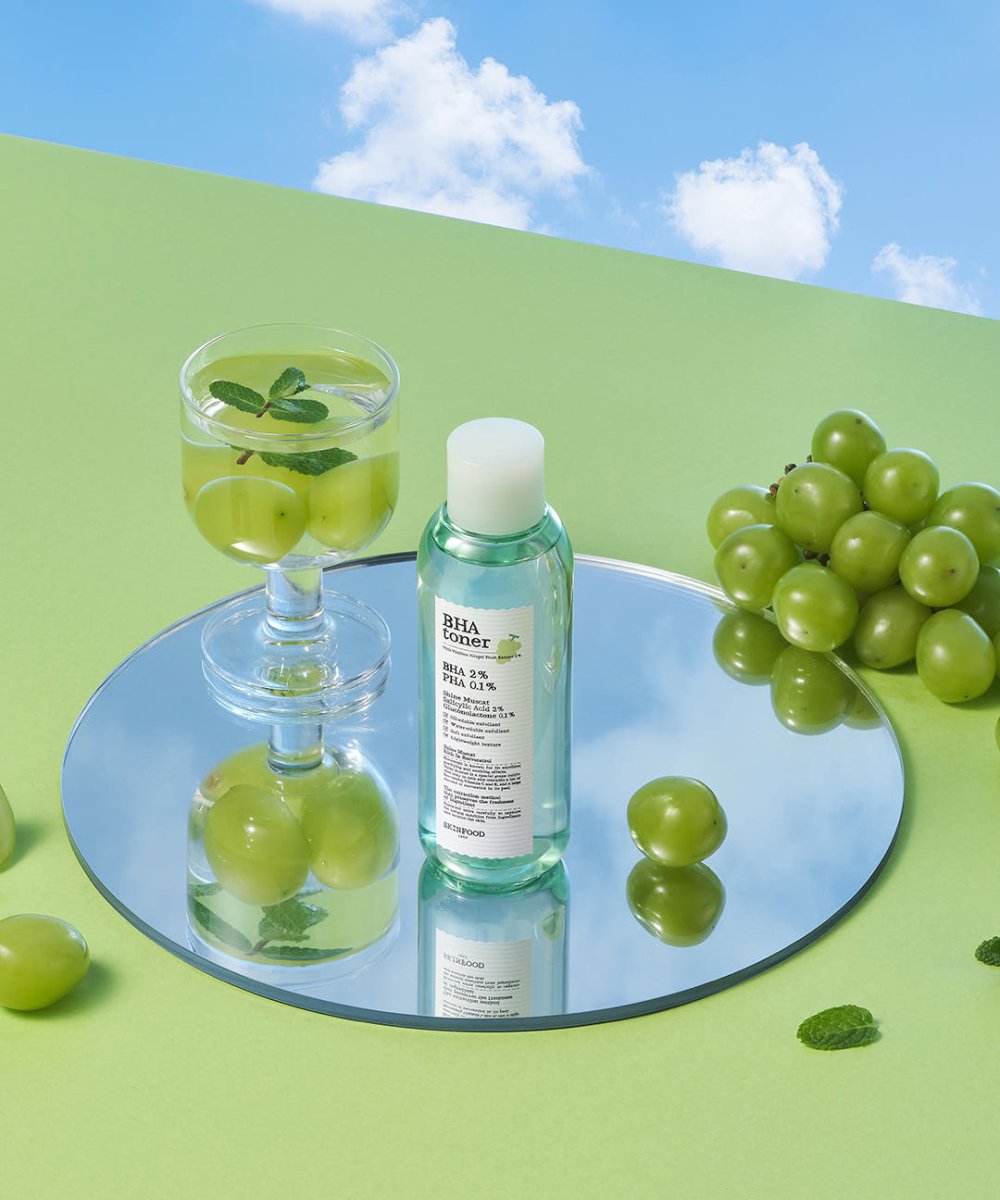 Clear green toner bottle on reflective surface surrounded by fresh muscat grapes and a glass with grape water infusion
