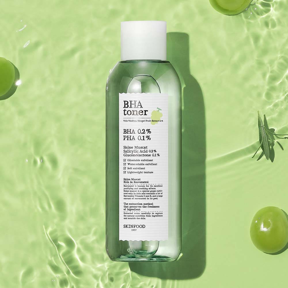 Clear green toner bottle with white label surrounded by Shine Muscat grapes and leaves on textured green surface