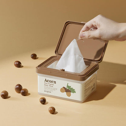 Hand pulling a single skincare sheet mask from a beige and brown container with acorn illustrations on light brown surface